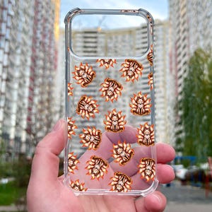 Pode incluir: Capa de telefone transparente com um padrão repetido de criaturas semelhantes a raposas, laranja e marrom. A capa é exibida em um fundo desfocado de edifícios e árvores, destacando os detalhes do design.
