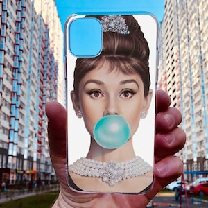 May include: A clear phone case with a white background featuring a woman with a pearl necklace and a large blue bubble gum. The woman has dark hair styled in a bun with a pearl headband.