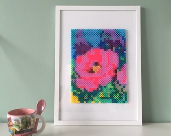 May include: A framed artwork featuring a colorful pixelated flower design. The flower is pink, blue, purple, and green. The artwork is framed in a white frame.