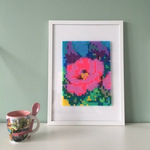 Op de afbeelding: Een ingelijst kunstwerk met een kleurrijk gepixeld bloeiende ontwerp. De bloem is roze, blauw, paars en groen. Het kunstwerk is ingelijst in een wit frame.
