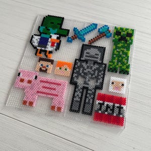 Op de afbeelding: Een verzameling pixel art figuren, waaronder een eend, een varken, een creeper, een skelet, een schaap en TNT, allemaal gemaakt van kleurrijke plastic kralen. Ook inbegrepen zijn twee karakterkoppen en een zwaard en pikhouweel.