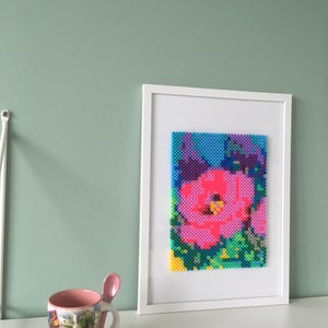 May include: A white framed picture with a colorful pixel art image of a pink flower. The flower is surrounded by green and blue pixels.