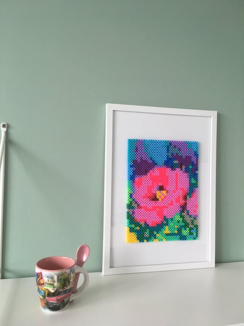 May include: A white framed picture with a colorful pixel art image of a pink flower. The flower is surrounded by green and blue pixels.