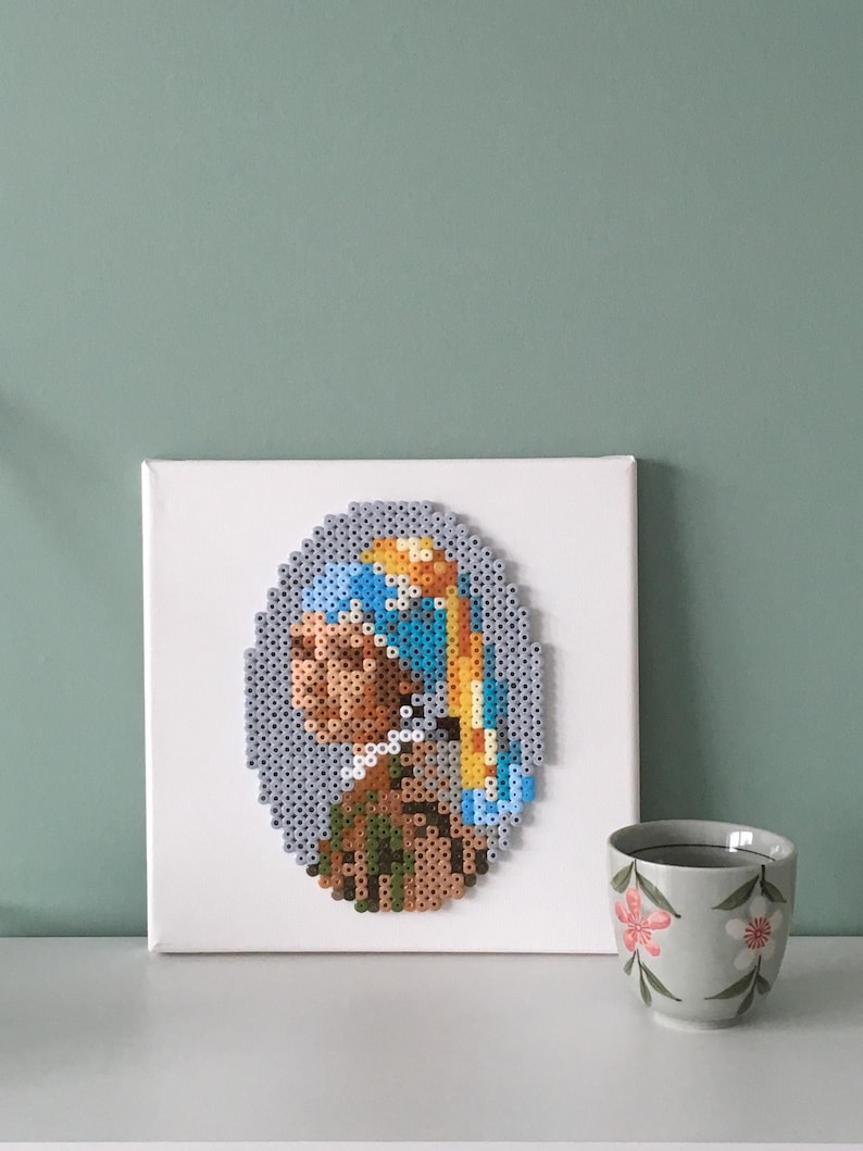 May include: A white canvas with a framed beaded artwork of a portrait of a woman with a blue and yellow headscarf and a pearl earring. The artwork is on a white shelf with a white teacup with a floral design.