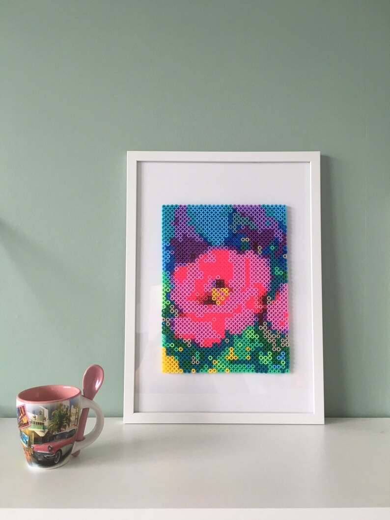 May include: A framed artwork featuring a colorful pixelated flower design. The flower is pink, blue, purple, and green. The artwork is framed in a white frame.