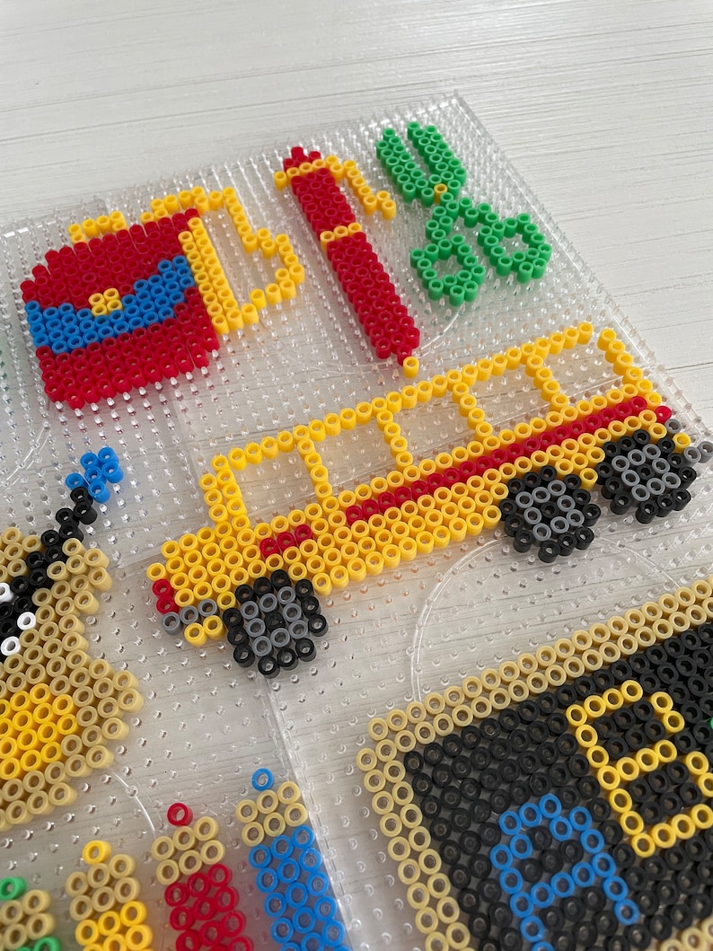 May include: A close-up of a colorful bead art project featuring a school bus, a fire extinguisher, scissors, and other school supplies. The beads are arranged in a grid pattern on a clear plastic board.