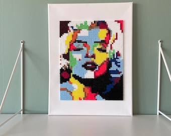 May include: A colorful beaded portrait of a woman with blue, yellow, red, white, and black beads. The portrait is framed and displayed on a white shelf.