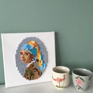 May include: A white canvas with a beaded portrait of a woman wearing a blue and yellow headscarf. The portrait is framed in a white oval. Two small teacups with floral designs are in the foreground.
