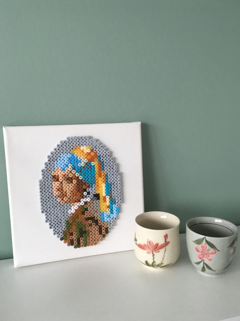 May include: A white canvas with a beaded portrait of a woman wearing a blue and yellow headscarf. The portrait is framed in a white oval. Two small teacups with floral designs are in the foreground.