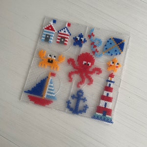 Op de afbeelding: Een set van 10 kleurrijke kralenontwerpen op een doorzichtige plastic plaat. De ontwerpen omvatten een zeilboot, een vuurtoren, een anker, een octopus, een krab, een vlieger en twee strandhuisjes.