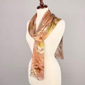 May include: A sheer, floral-patterned scarf in shades of brown, tan, and yellow is draped over a mannequin. The scarf's design features a delicate floral print, adding a touch of elegance. The scarf is lightweight and appears to be made of a soft, flowing fabric.