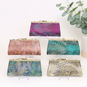 Könnte beinhalten: Fünf kleine, rechteckige, goldumrandete Clutch-Taschen mit verschiedenen Pfauenfedermustern. Die Clutch-Taschen sind in einer Reihe auf einer weißen Oberfläche angeordnet. Die Muster umfassen Rosa, Orange, Türkis, Blau und Grau.