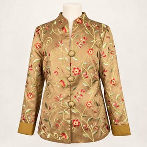 May include: A tan jacket featuring a mandarin collar and long sleeves with wide cuffs. The garment is decorated with an embroidered pattern of red flowers and green vines, and it fastens down the center with three round fabric-covered buttons.