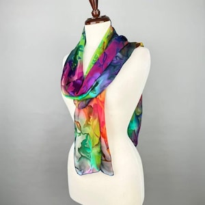 May include: A lightweight scarf featuring a vibrant, multi-colored abstract floral pattern in shades of magenta, lime green, bright orange, and deep purple. The scarf is draped around a white mannequin torso, displaying a fluid, semi-sheer fabric texture.