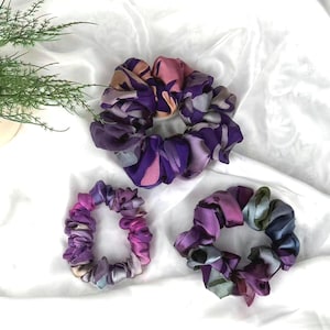 May include: Three scrunchies in shades of purple, pink, and gray. The scrunchies have a patterned design and are made of a soft, silky material. The scrunchies are arranged on a white background.