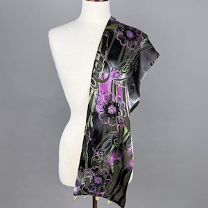 May include: A patterned scarf in black, purple, and green is displayed on a mannequin. The scarf has a floral design with swirls and gold-coloured beads at the bottom. The fabric appears to be silky and lightweight, suitable for draping.