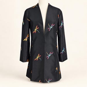 May include: A black open-front jacket featuring long sleeves and a v-neckline. The garment is decorated with an all-over embroidered pattern of dragonflies in vibrant shades of blue, yellow, orange, and magenta scattered across the fabric.