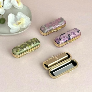 Puede incluir: Cuatro pequeñas cajas decorativas con motivos florales. Las cajas están hechas de metal dorado y tienen una tapa con bisagras. Dos de las cajas son verdes con detalles dorados, una es rosa con detalles negros y una es morada con detalles negros. Las cajas están todas cerradas.