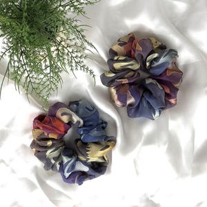 May include: Two scrunchies with a colorful abstract pattern in shades of purple, blue, orange, and yellow. The scrunchies are made of a silky fabric and are displayed on a white, wrinkled surface.