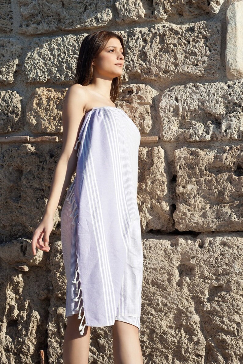 May include: A woman wearing a white and light purple striped towel with fringe. The towel is wrapped around her body like a dress. She is standing in front of a stone wall.
