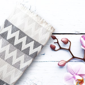 May include: A white and gray patterned throw blanket with a chevron design, lying on a white wooden surface with pink orchid flowers.
