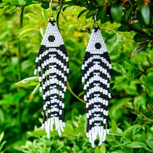 Puede incluir: Un par de pendientes de cuentas con un diseño de rayas blancas y negras, colgando de una rama verde y frondosa. Los pendientes tienen forma de lágrima con flecos blancos en la parte inferior. El texto "Dragonfly Earrings" y "ART FOR YOUR EARS" es visible.