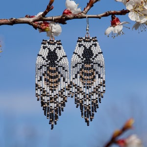 Puede incluir: Un par de pendientes de libélula con cuentas en negro, blanco y dorado, con flecos. Los pendientes cuelgan de una rama con flores blancas sobre un cielo azul. El texto "Dragonfly Earrings, Art for your Ears" está presente en la imagen.