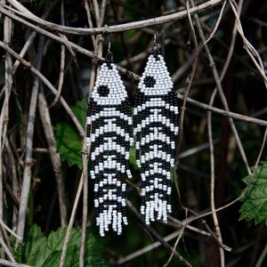 以下が含まれることがあります： 黒と白のビーズで作られた、魚の骨のデザインのビーズイヤリング。 イヤリングは、小枝と葉を背景に、シルバーのフックから吊り下げられています。「Dragonfly Earrings」と「Art for your ears」の文字が見えます。