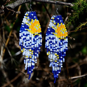 May include: A pair of long, dangling beaded earrings featuring a vibrant pattern of blue, white, and yellow glass beads. The design incorporates a circular yellow sun motif against a blue and white background, with fringe-style beaded strands.