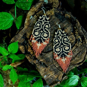 May include: A pair of long, beaded earrings with a geometric pattern in black, gold, and copper. The earrings are hanging from a wooden surface.
