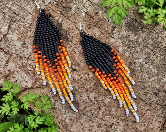 May include: A pair of beaded earrings with a black, orange, yellow, and white color scheme. The earrings have a fringe design and are displayed on a textured, brown surface with green foliage.