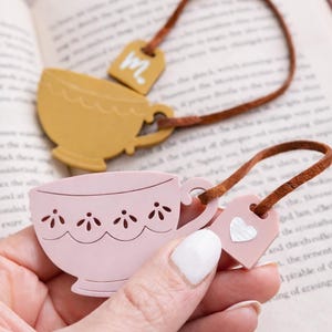 May include: Pink and yellow teacup-shaped bookmarks with brown leather cords. One bookmark has a heart-shaped tag, the other has a tag with the letter "M". The bookmarks are on an open book.