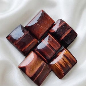 May include: Five polished, rectangular red tiger's eye gemstones arranged in a cluster. Each stone features a smooth, glossy surface with distinct chatoyant bands of deep mahogany, rust, and golden-brown hues that shimmer under the light.