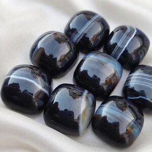 May include: A collection of eight polished, rounded square stones featuring a deep black base color with distinct, horizontal white and translucent gray banding patterns throughout each individual piece, showcasing a smooth and reflective surface.