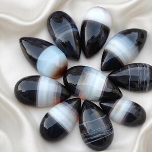 May include: A collection of nine polished, teardrop-shaped black and white banded agate gemstones arranged in a circular pattern. Each stone features smooth, glossy surfaces with distinct horizontal white stripes contrasting against deep black sections.