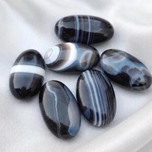 May include: A collection of six polished, oval-shaped stones featuring smooth surfaces. The stones are primarily black with distinct white and gray banded patterns, swirls, and stripes. Each stone has a glossy finish and unique natural markings.