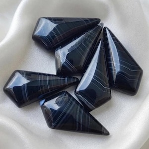May include: A collection of five polished, shield-shaped black agate stones featuring distinct, thin white horizontal banding. Each stone has a smooth, glossy surface with a reflective finish, showcasing natural layered patterns in a dark color palette.