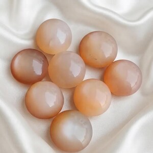 May include: A collection of eight smooth, round, polished stones in varying shades of soft peach and translucent light orange. Each stone features a glossy, reflective surface with subtle internal variations, arranged in a cluster.