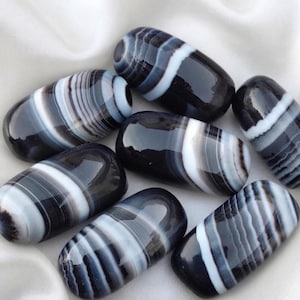 Puede incluir: Un conjunto de siete piedras pulidas de forma ovalada que presentan bandas horizontales distintivas en tonos negro, gris oscuro y blanco. Cada piedra tiene una superficie lisa y brillante con patrones variados de rayas blancas opacas sobre tonos oscuros.