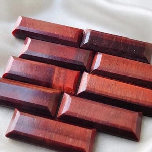 May include: A collection of seven rectangular, faceted red tiger's eye gemstones. Each stone features a polished surface with deep reddish-brown tones and a distinct chatoyant luster, arranged in a staggered, overlapping pattern on a smooth surface.