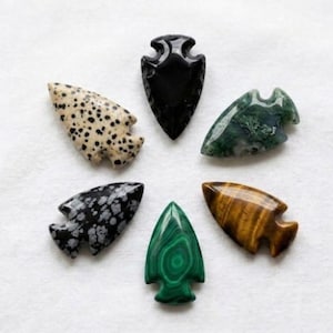 Può includere: Sei punte di freccia in pietra levigata disposte in cerchio. La collezione comprende un'ossidiana nera solida, una pietra color crema con macchie nere, un'agata muschiata verde, un'ossidiana fiocco di neve bianca e nera, una malachite verde e un occhio di tigre dorato.