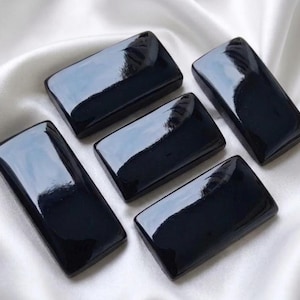 Peut inclure: Cinq cabochons rectangulaires noirs avec une surface lisse, polie et réfléchissante. Chaque pièce présente des bords arrondis et une finition brillante et plate qui capte la lumière, créant une apparence uniforme pour les cinq objets.