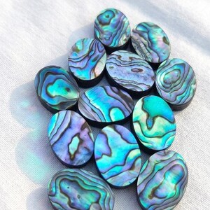 May include: A collection of oval-shaped abalone shell cabochons. The shells display iridescent swirls of blue, green, and purple hues. The cabochons are arranged on a white fabric surface.