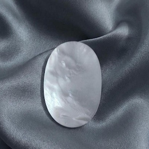 May include: An oval-shaped piece of iridescent mother-of-pearl with a smooth, polished surface. The object displays a natural, swirling pattern of white and soft gray tones, reflecting light with a subtle, pearlescent sheen across its flat face.