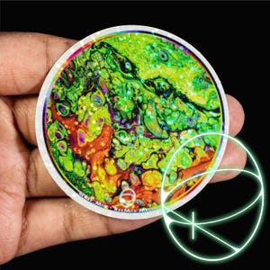 May include: A round sticker with a vibrant, abstract design in shades of green, yellow, orange, and purple. The sticker has a white border and the text "@Kee-Mix - KeeMix Media" is printed on the bottom. A glowing neon green graphic is in the lower right.