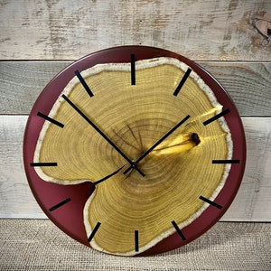Puede incluir: Un reloj redondo de madera con un borde de resina roja. La esfera del reloj está hecha de una rodaja de madera natural con anillos de crecimiento visibles. Se ven las manecillas negras de las horas y los minutos.
