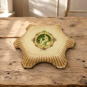 May include: A hexagonal ceramic trivet with a cream-coloured centre featuring a green cameo-style illustration of a seated figure and a cherub, surrounded by a floral wreath. The edges are scalloped, ribbed, and finished in a light brown glaze.