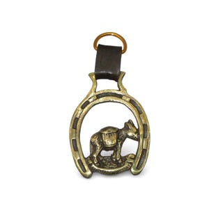 Könnte beinhalten: Ein antikes Pferdegeschirr aus Messing in Hufeisenform mit einem detaillierten Relief eines Esels. Es hat einen dunkelbraunen Lederriemen und einen Messingring. Der Esel trägt einen Sattel. Ein dekoratives Element.
