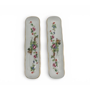 May include: Two long, rectangular white porcelain serving dishes with rounded corners and gold-trimmed edges. Each dish features a central hand-painted floral motif in shades of pink, blue, and green, accented with small brown architectural details.