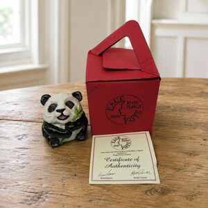 May include: A ceramic panda figurine holding a green bamboo stalk, accompanied by a red cardboard box with a handle and a printed Certificate of Authenticity. The box features a black circular logo reading Face Kevin Francis Pots 2000.